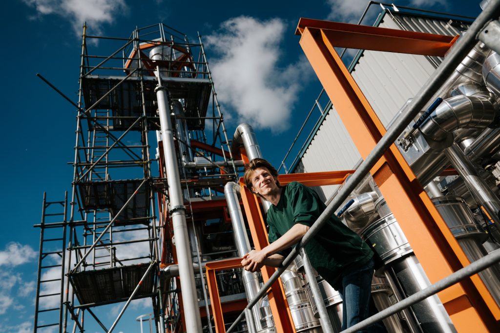 Een industriële omgeving met steigers en metalen buizen. Een persoon leunt op een reling.